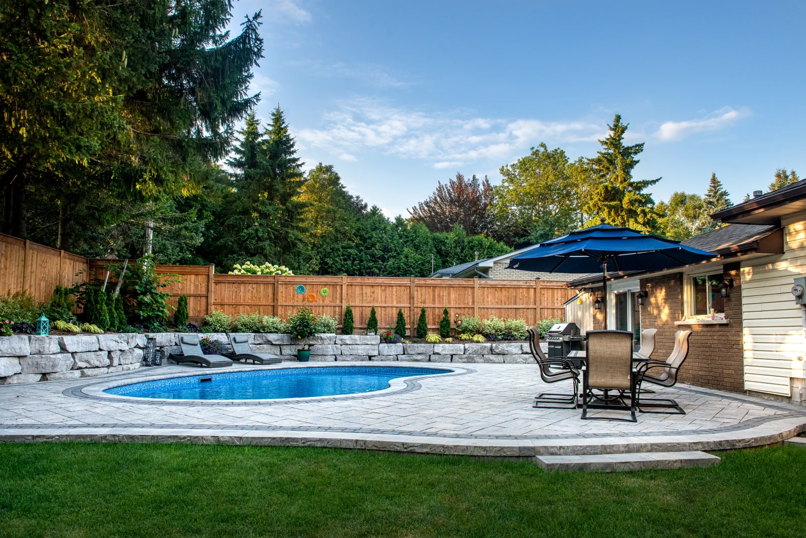 Wide ground-level view of a curved backyard pool, interlock patio and armour stone retaining wall in Ancaster.