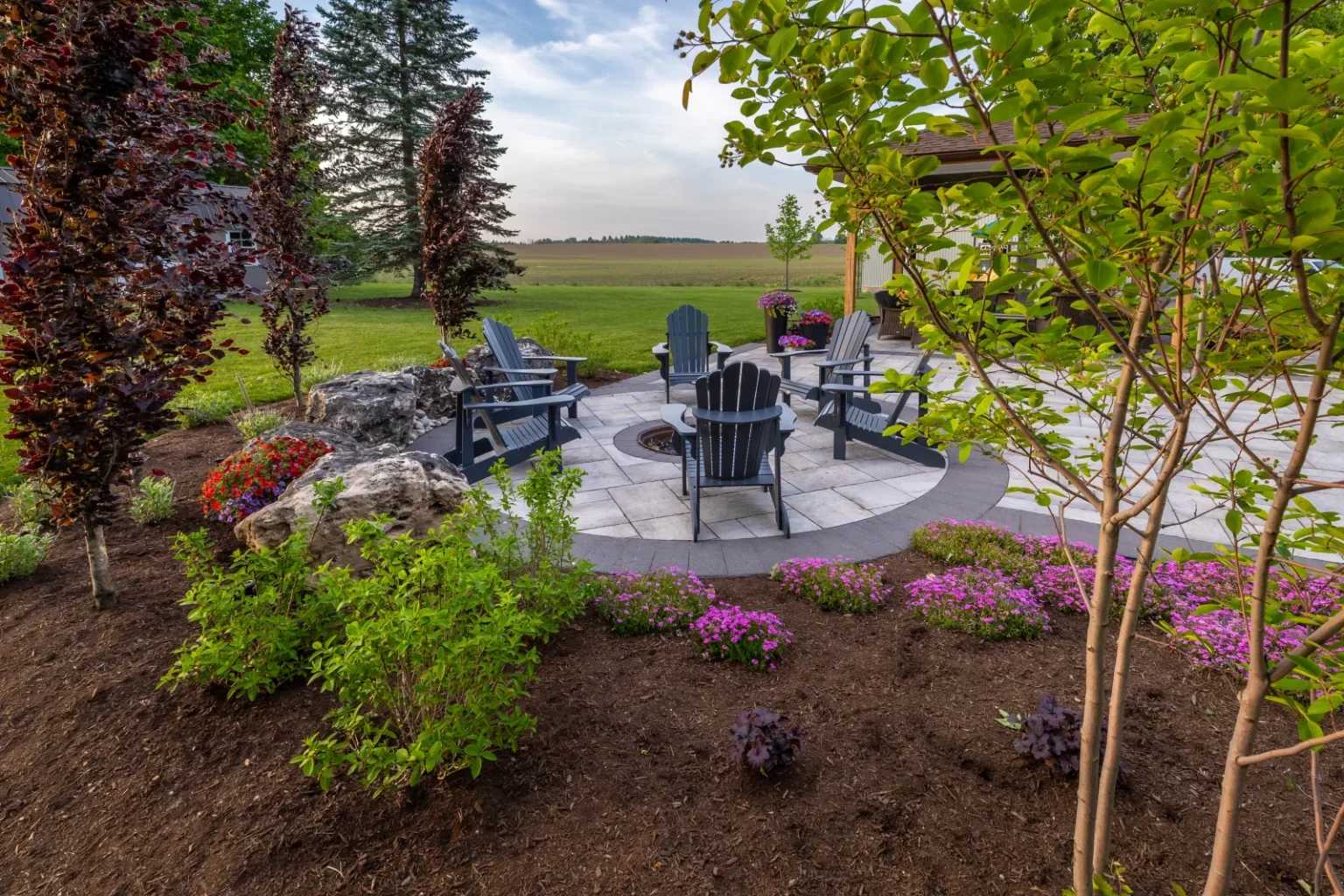 Alternate view of the circular fire-pit patio framed by flowering plants and young trees in Flamborough.