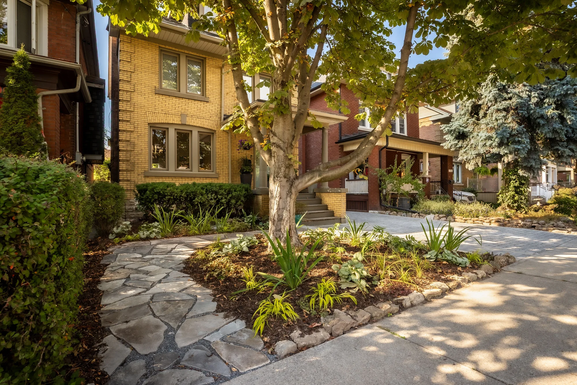 A flagstone entrance path and planted front garden beneath a mature shade tree at a brick home in Hamilton.