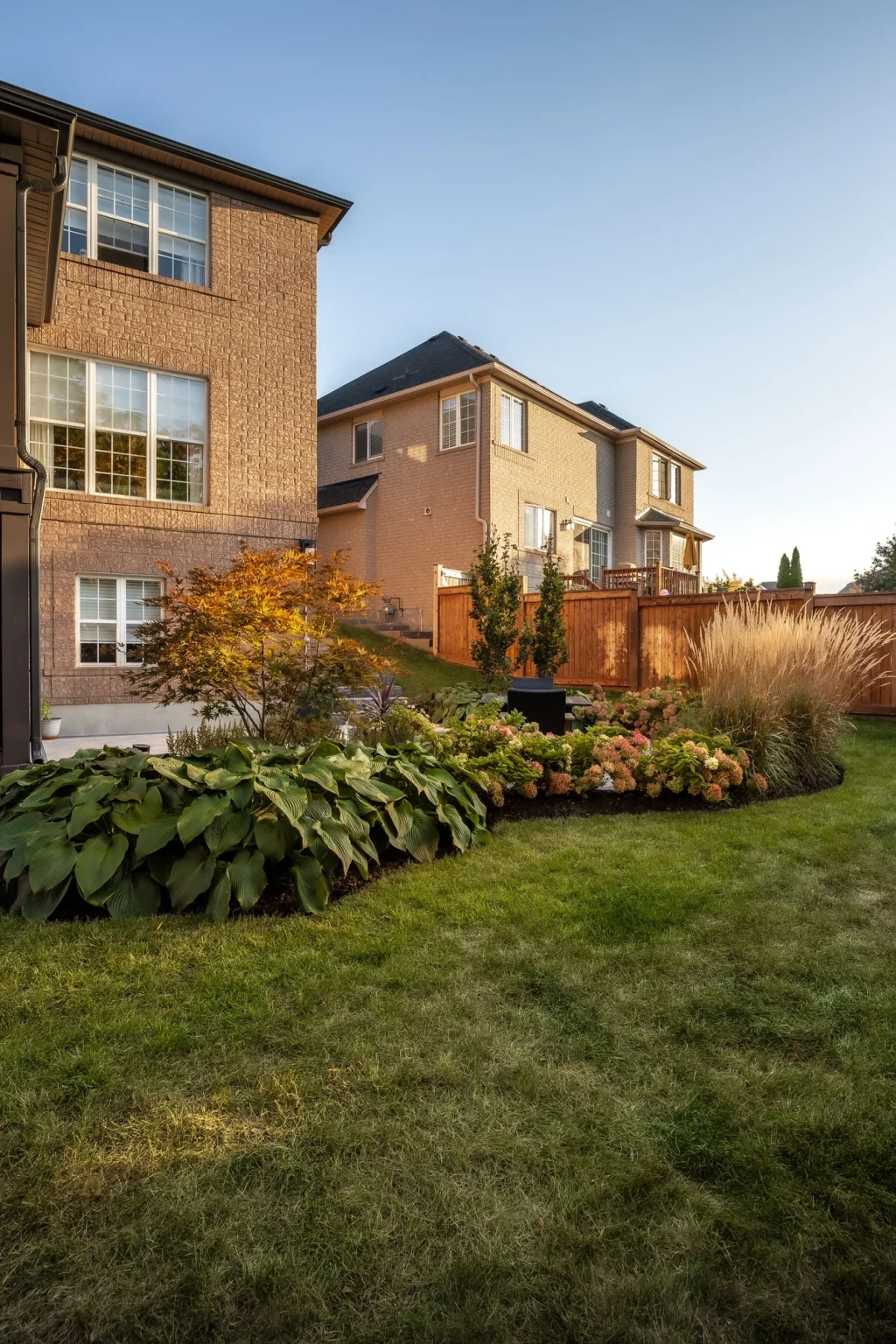 Wide lawn-facing view of a brick house, raised deck, planting beds and the patio extending along the rear of the home in Oakville.
