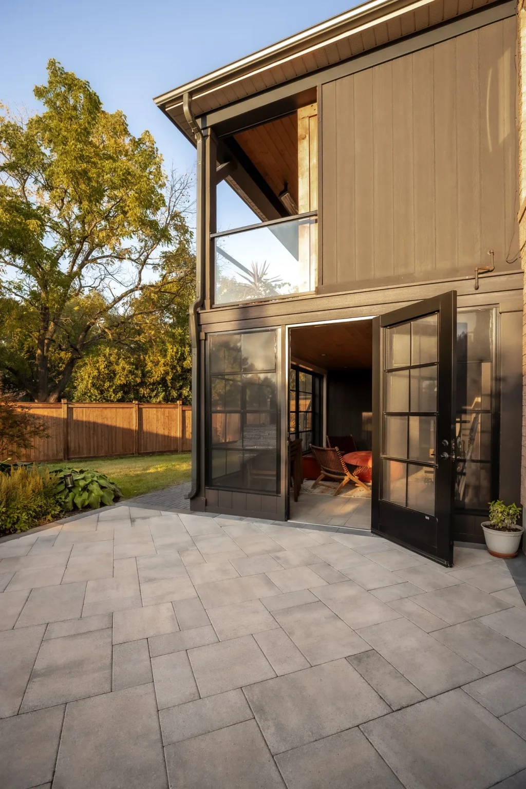 Portrait photograph of the house-facing grey stone patio beneath a raised covered deck with wood-clad ceiling, dark railings and glass doors in Oakville.