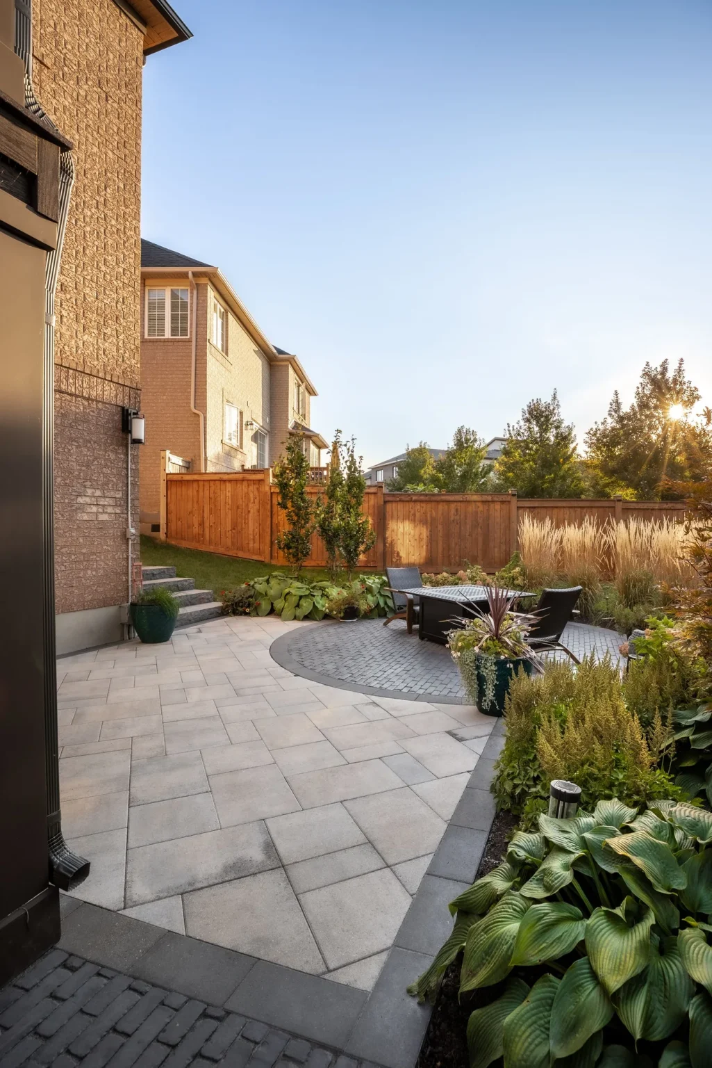 Portrait view along a light-grey stone patio with curving borders, lush hostas, ornamental grasses, flowering beds and timber fencing in Oakville.