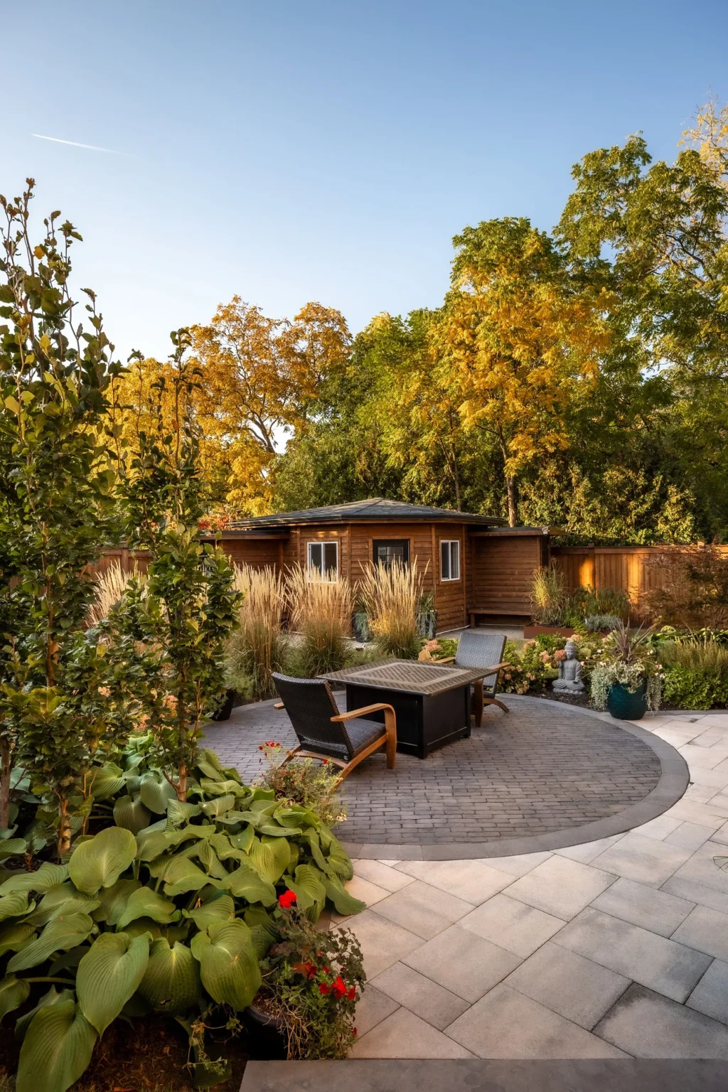 Portrait view through colourful planting toward the circular interlock patio, rectangular outdoor fire table and ornamental grasses in Oakville.