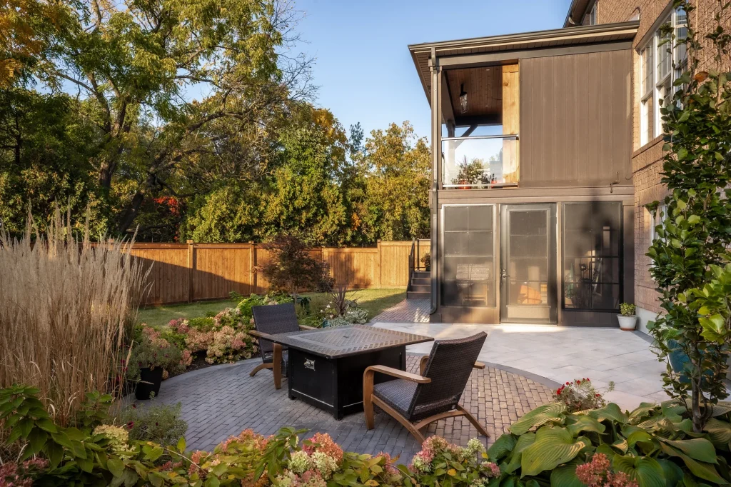 A stone patio with a fire table and seating surrounded by planted borders beside an Oakville home.