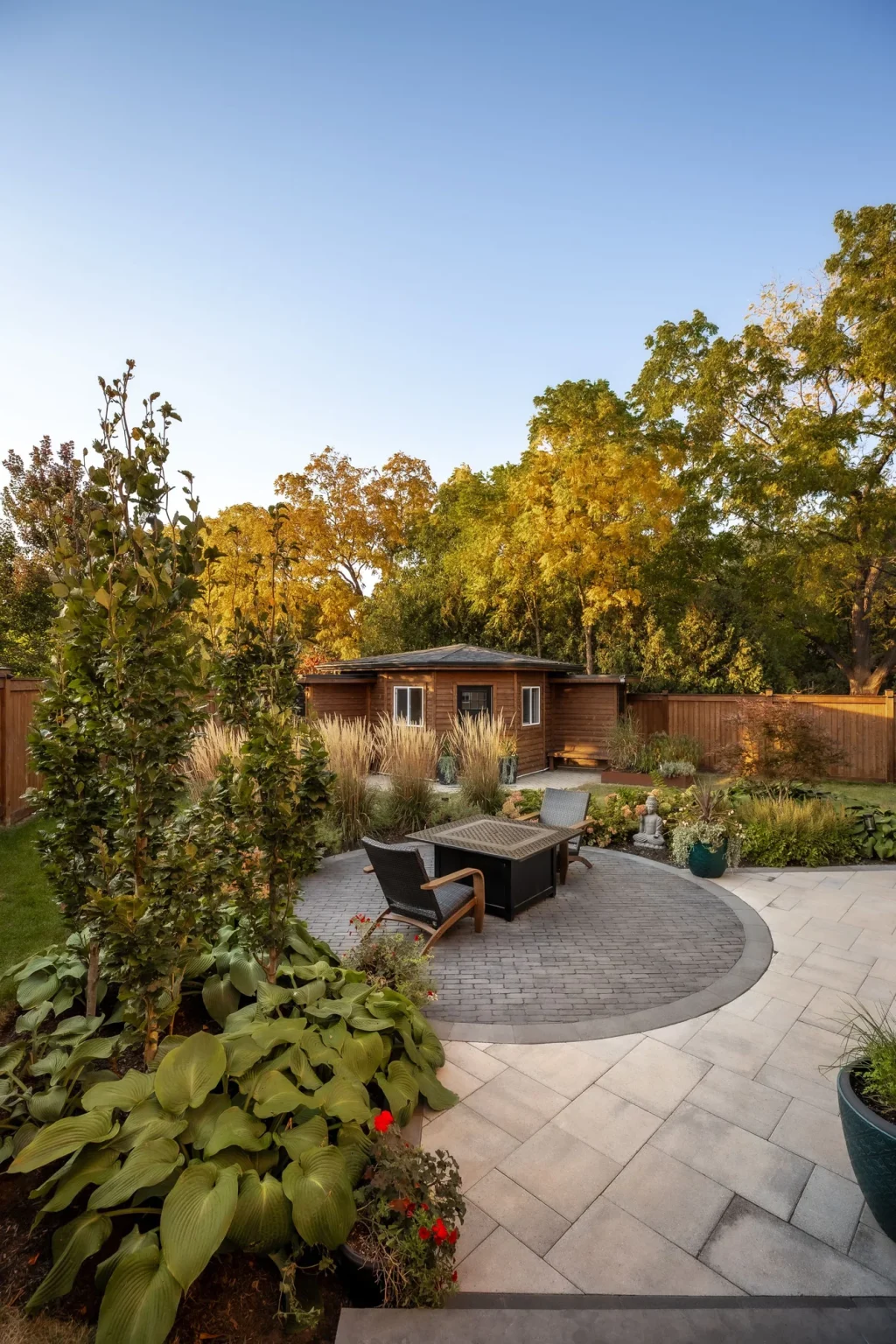 Portrait garden-level view of rounded stone patio seating around a rectangular fire table enclosed by flowering plants, shrubs and mature trees in Oakville.