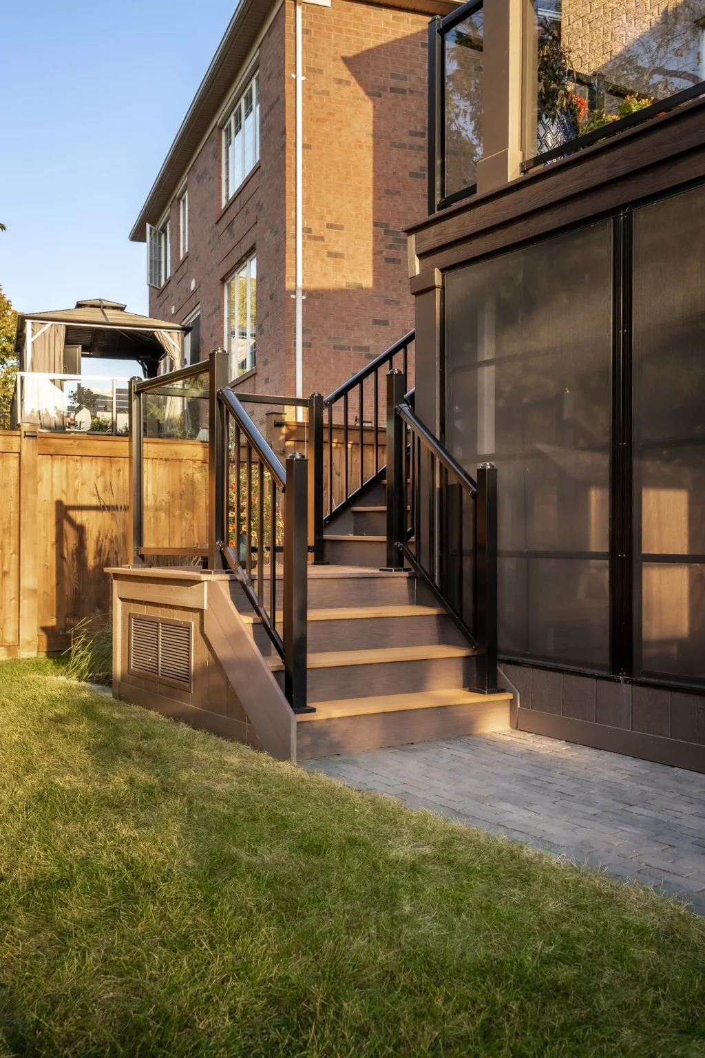 Portrait view of steps with black metal railings beside a raised outdoor deck and the lower stone patio at a brick house in Oakville.