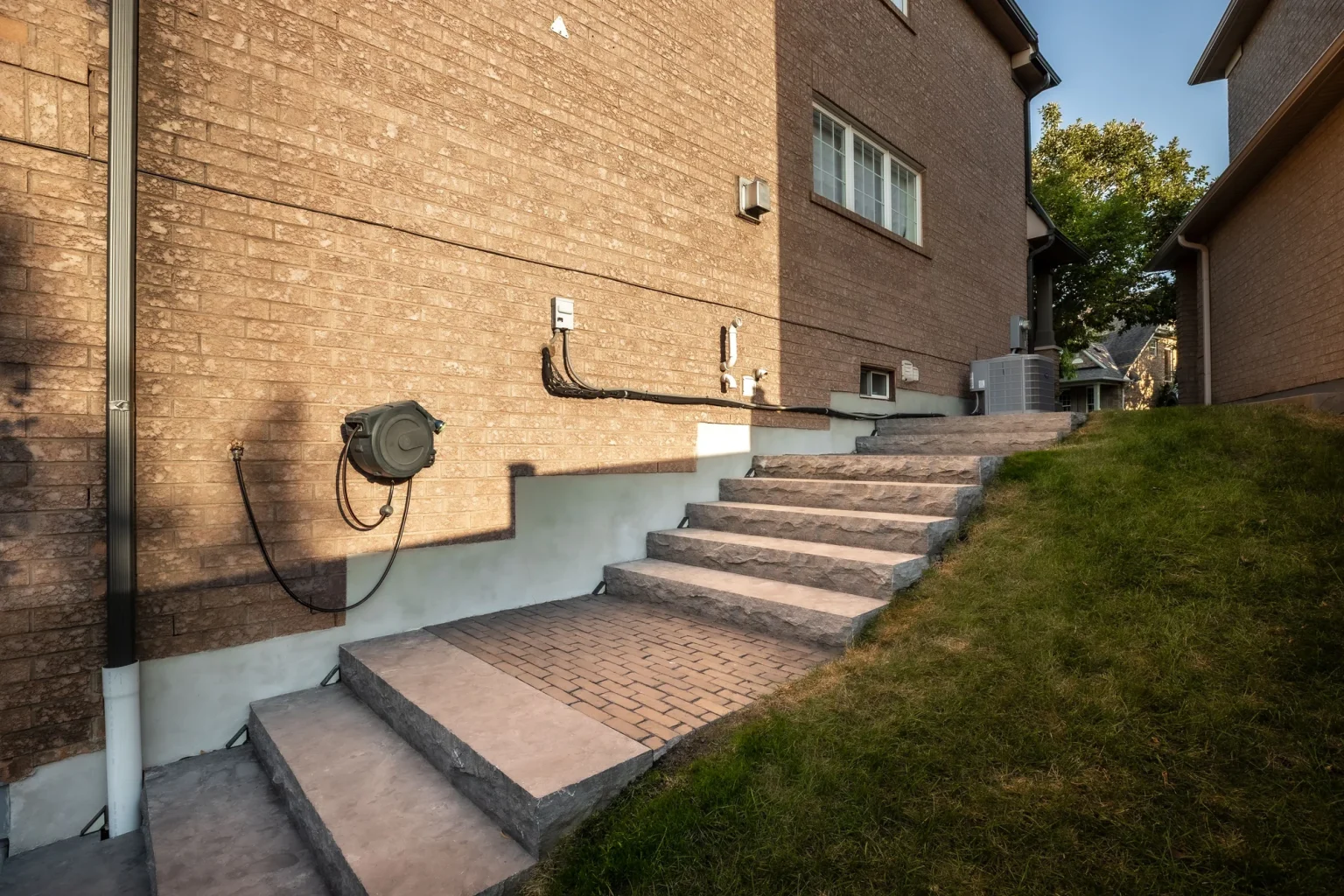 Side-of-house view of wide grey steps climbing between a brick wall and grass slope from a compact patio in Oakville.