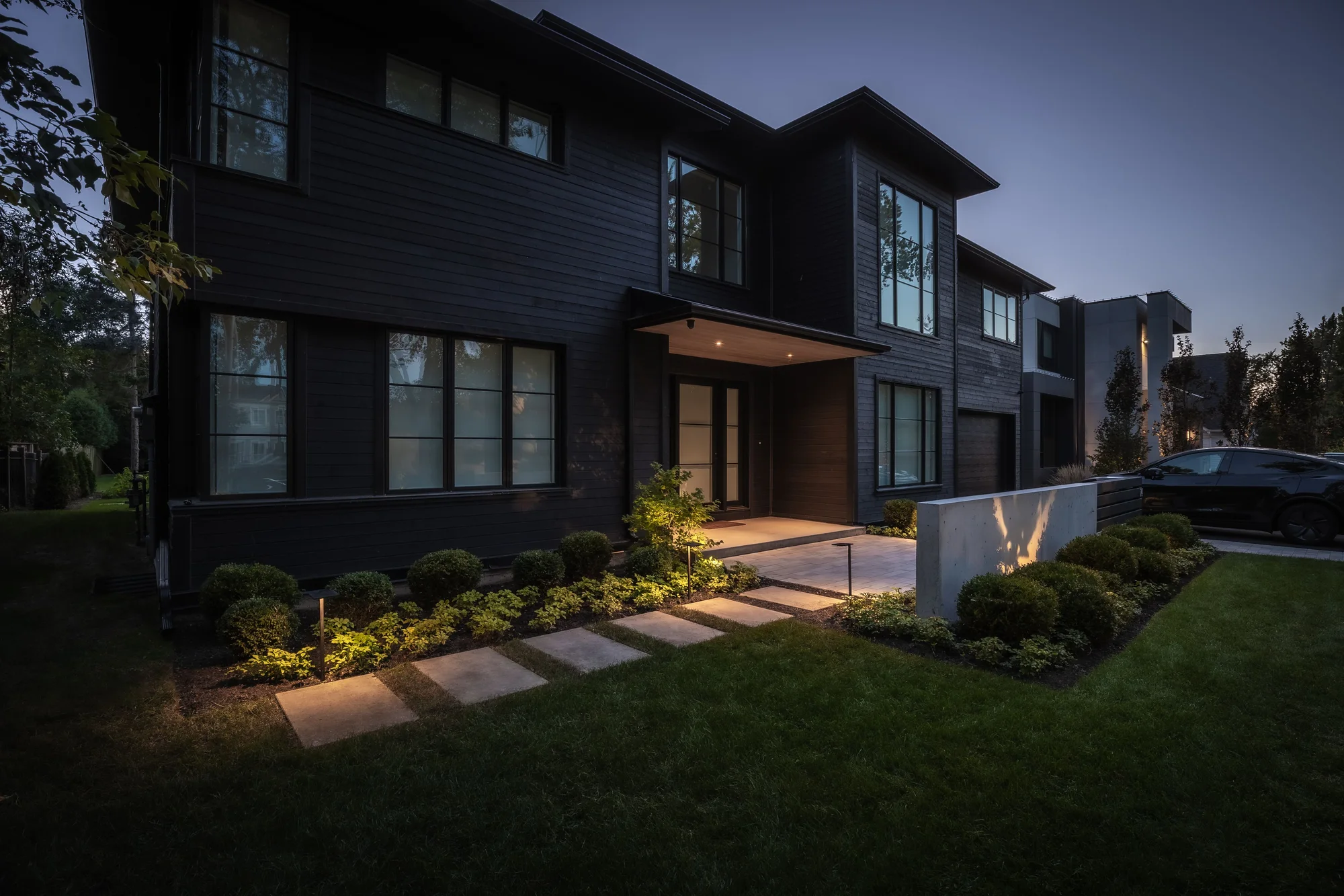 A modern black home at dusk with illuminated planting and concrete stepping stones across the lawn in Oakville.